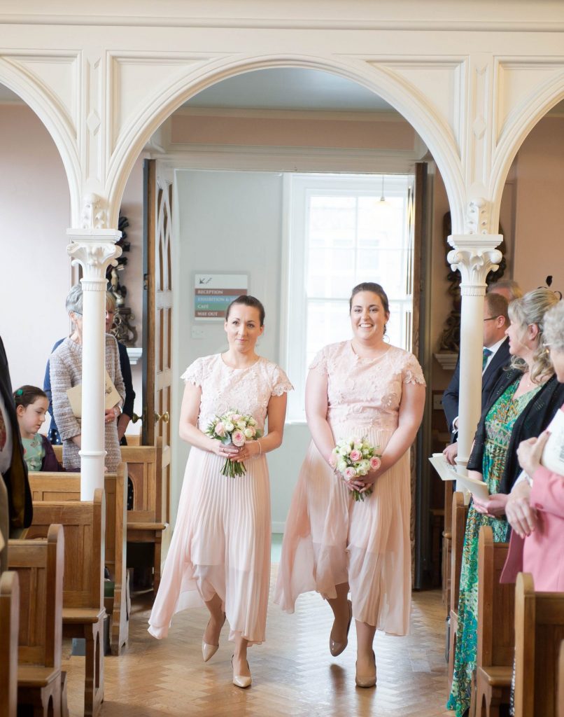 Two bridesmaids in dusky pink lace dresses holding pink and ivory wedding posies walk up the aisle at the Bar Convent in York, North Yorkshire