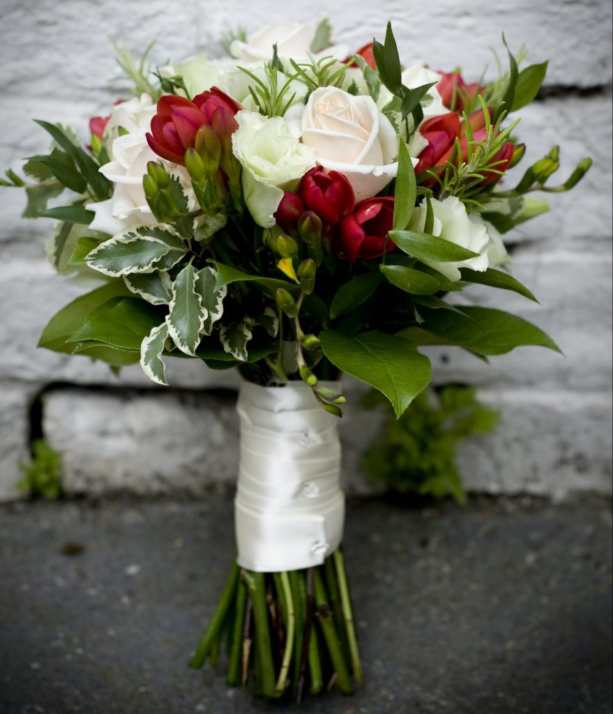 Handtied bridal bouquet of cream roses, red freesia, lisianthus, dark green foliage and variegated foliage finished off with satin ribbon and diamante pins.